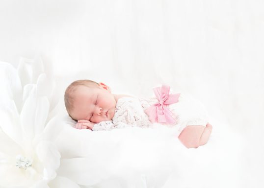 Schlafendes Baby in weißer Decke mit rosa Schleife und Blume im Hintergrund.